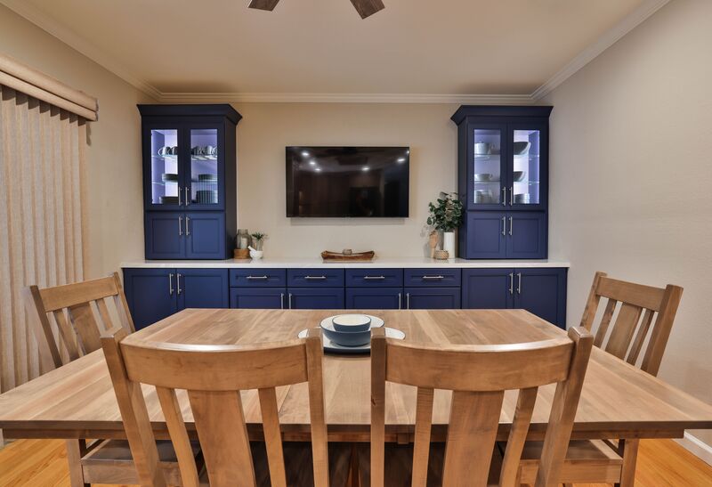 Wooden dinning table and chairs with a tv in the middle of blue cabinets topped with a white counter top 