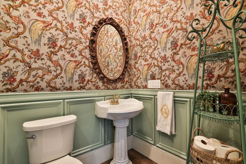 small white sink with oval mirror and red and off-white bird patterned wallpaper with square light green wood paneling. 