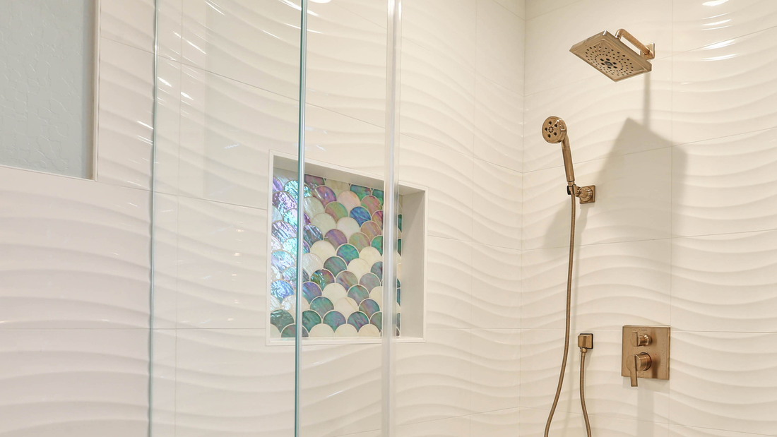 White texual field tile with a wave design are offset by a shower niche with colorful mermaid tiles and brass fixtures