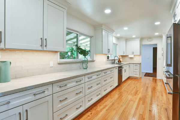 Long kitchen with white cabinets and backsplash and light hardwood floor.